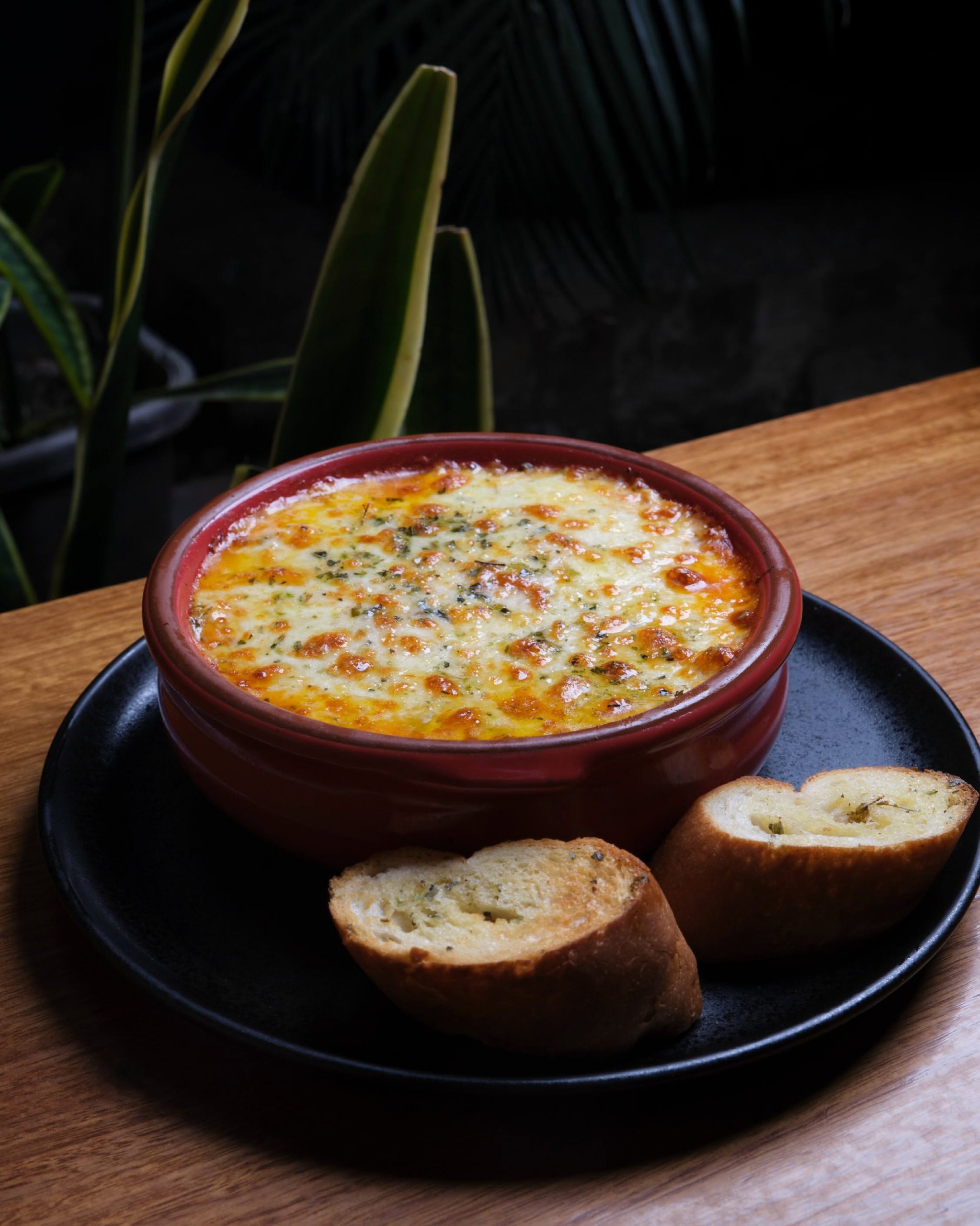 Cazuela de lasaña mixta recién horneada con queso gratinado y pan de ajo sobre mesa de madera, Morelia Parrilla Loja