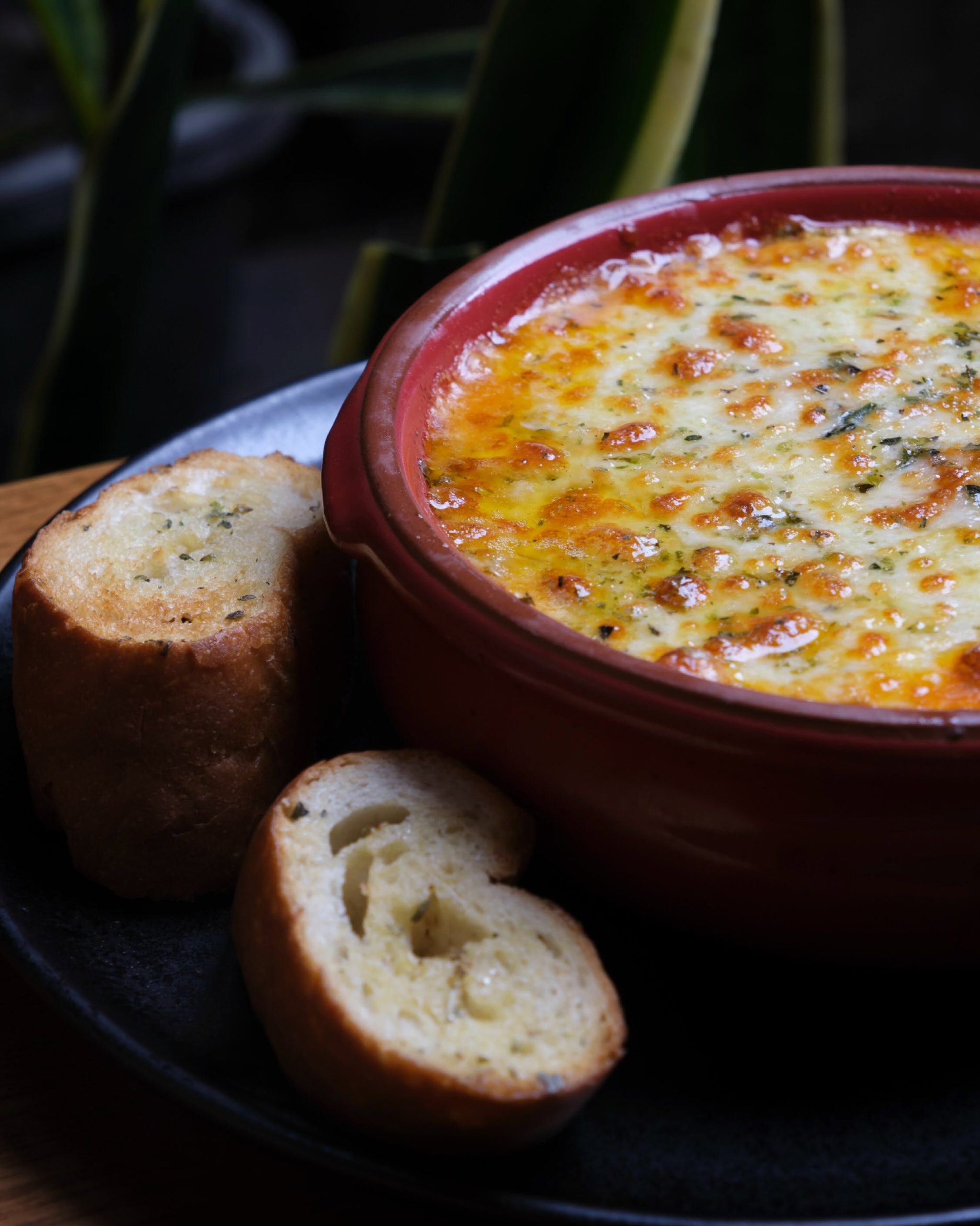 Primer plano de lasaña mixta gratinada en cazuela de barro roja junto a pan de ajo tostado, Morelia Parrilla Loja